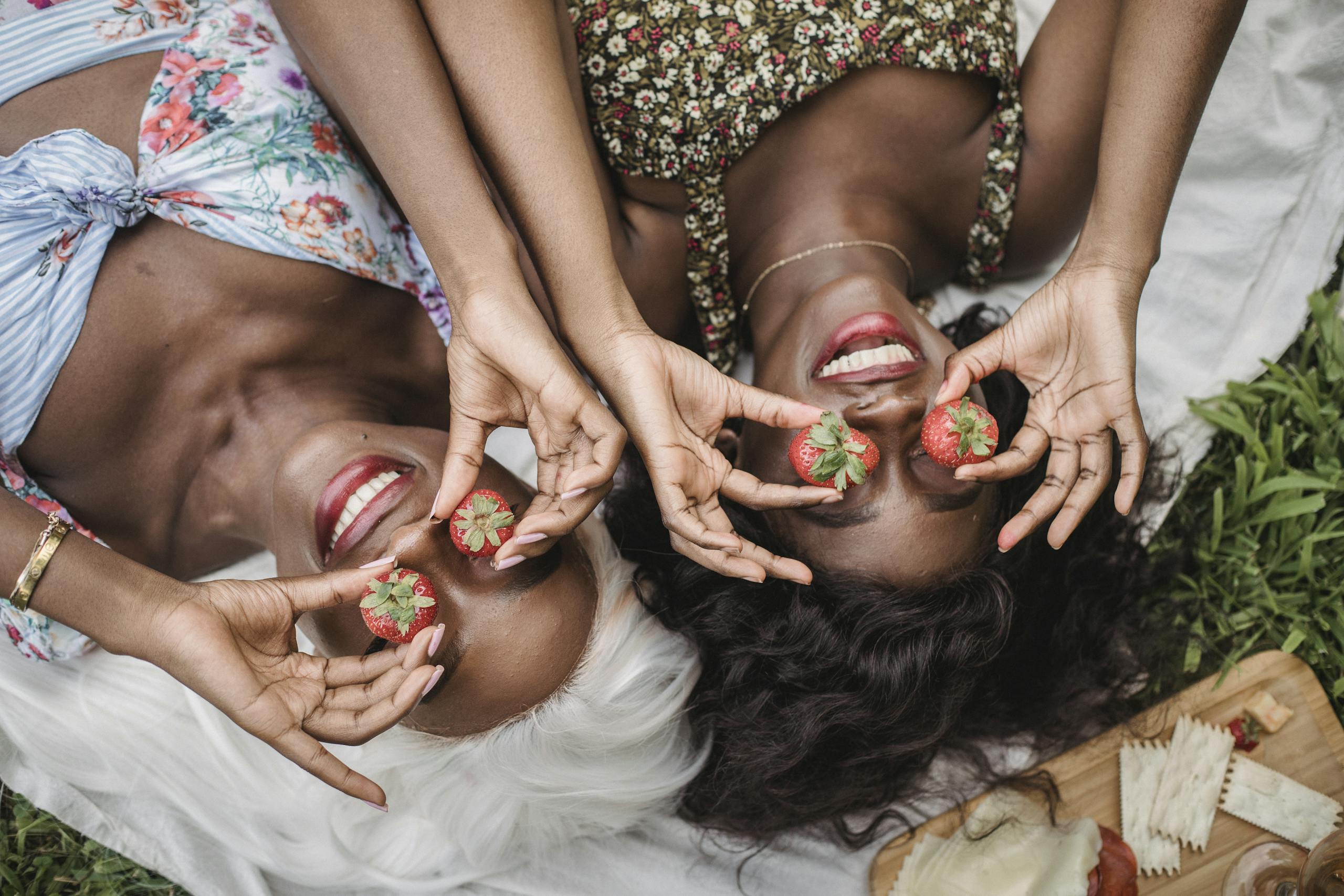Two women lying on a picnic blanket, laughing while holding strawberries over their eyes.